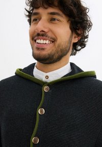 Uomo sorridente con capelli ricci che indossa un maglione scuro di lana con bottoni e rifiniture verdi sopra una camicia bianca.