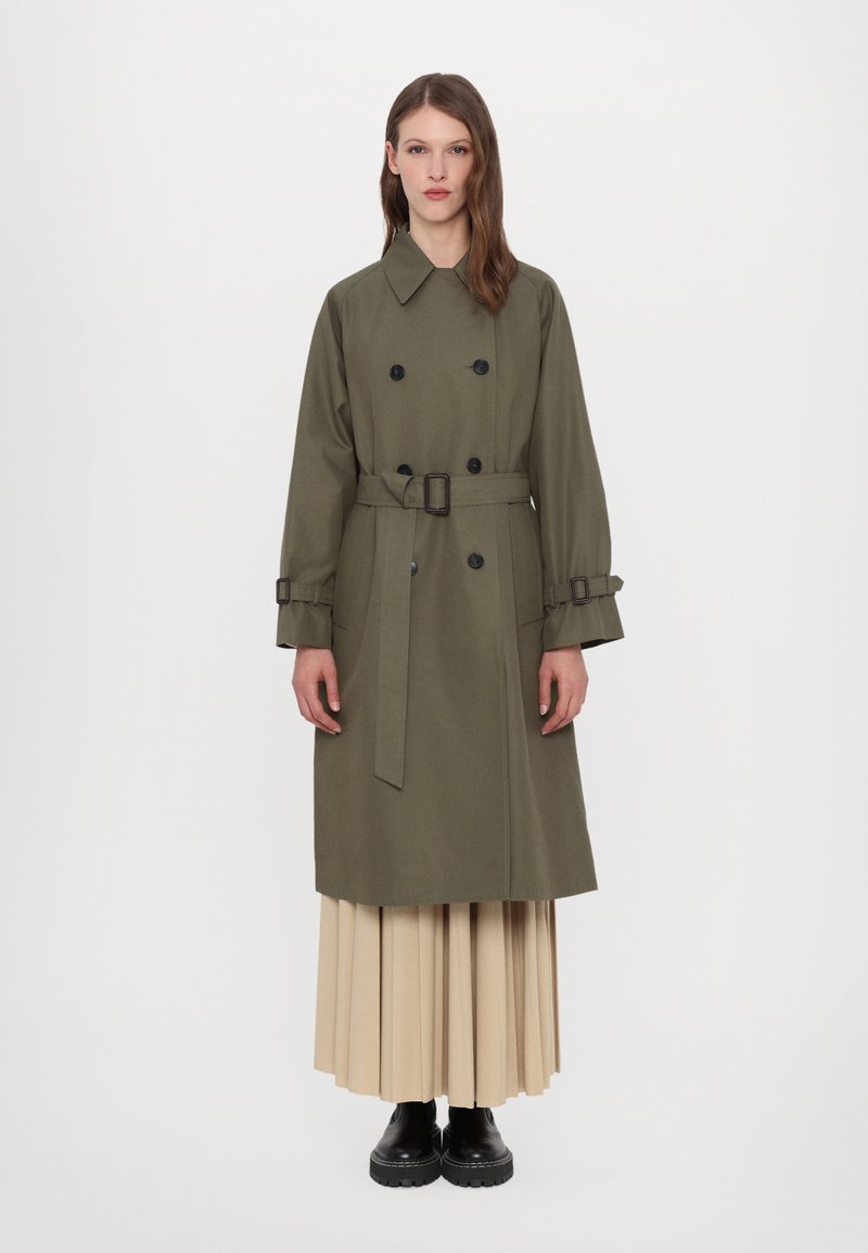 Olíva zöld trench coat dupla mellső résszel, illeszkedő övvel és fekete gombokkal. Párosítsd bézs redős szoknyával és fekete bokacsizmával.
