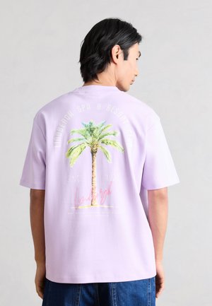 Mężczyzna noszący jasno-fioletową koszulkę z dużą zieloną grafiką palmy i różowym tekstem na plecach.