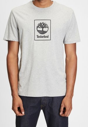 Grå bomulds t-shirt med et sort Timberland-logo med et trædesign, rund halsudskæring og korte ærmer. Sammen med mørkeblå jeans.