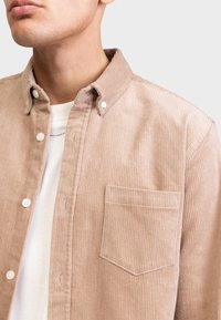 Beige corduroy overhemd met een button-down kraag, textuurstof, voorzak en witte knopen over een wit t-shirt.