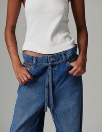 Άτομο που φοράει φαρδιά μπλε τζιν με ζώνη δεμένη από τζιν και λευκό ριμπ τοπ χωρίς μανίκια, με τα χέρια σταθερά στις τσέπες του τζιν.