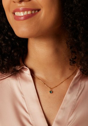 Goldene Kettenhalskette mit einem kleinen blauen Edelsteinanhänger. Die Trägerin hat lockige Haare und trägt eine blassrosa Satinbluse.