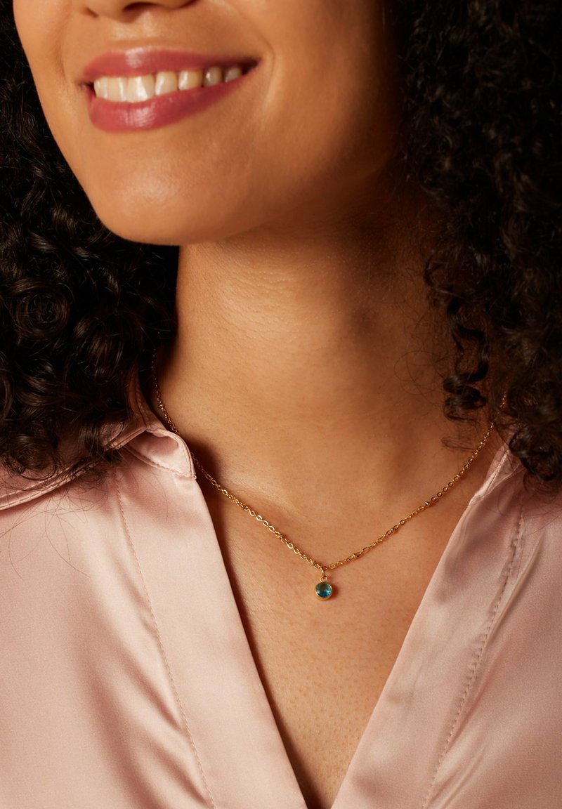 Gouden ketting met een kleine blauwe edelsteen hanger. De drager heeft krullend haar en is gekleed in een lichtroze satijnen blouse.