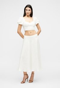Femme portant un crop top en dentelle florale blanche à manches bouffantes, une jupe midi blanche et des sandales marron à talons hauts, debout les mains derrière le dos.