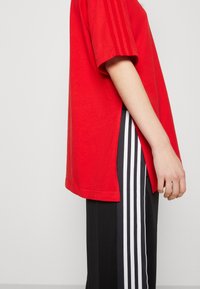 Červené bavlněné tričko s krátkým rukávem a bočními rozparky, spárované s černými kalhotami s třemi bílými pruhy na bocích.