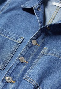 Gros plan d'une veste en jean bleue avec des boutons-pression argentés et deux poches poitrine avec des coutures jaunes.