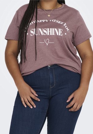 Mujer con camiseta color malva con el texto "Tan feliz de verte, rayito de sol" y jeans azul oscuro, complementada con collares dorados.