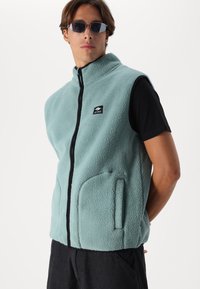 Vesta din fleece de culoare teal deschis, cu guler înalt, fermoar frontal și buzunare laterale. Caracterizată printr-un mic logo negru pe piept.