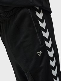 Schwarze Sporthose mit weißen Chevron-Streifen an der Seite, einer Reißverschlusstasche und einem kleinen Bienenlogo neben der Tasche.