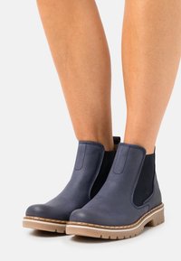 Stivali alla caviglia in pelle blu scuro con lati elasticizzati e suola in gomma texturizzata color tan. Presentano una punta arrotondata e dettagli di cucitura rinforzata.