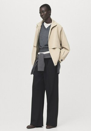 Wysoka osoba ubrana w beżową kurtkę na szarym swetrze i czarne, szerokie spodnie, stoi z rękami w kieszeniach na białym tle.