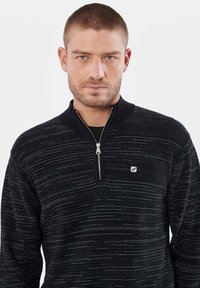 Pull noir avec un col zippé, présentant un motif texturé avec de subtiles lignes grises. Inclut un petit logo sur la poitrine.