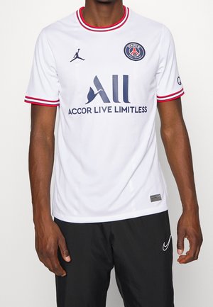 Uomo che indossa la maglia bianca del Paris Saint-Germain con bordi rossi e pantaloni neri, in piedi su uno sfondo semplice.