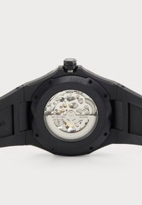 Schwarze Silikonarmbanduhr mit einem transparenten Gehäuseboden, der ein silbernes Miyota-Uhrwerk und verschiedene mechanische Komponenten zeigt.