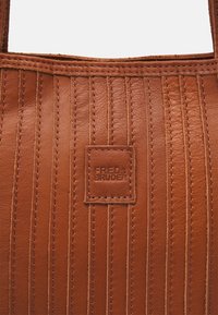 Hnědá kožená taška přes rameno s vertikálním prošitím, texturovaným povrchem a obdélníkovou vyraženou logovou nášivkou.