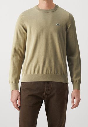 Homme portant un pull vert olive à col rond avec un petit logo crocodile sur la poitrine et un pantalon marron foncé, debout devant un fond uni.