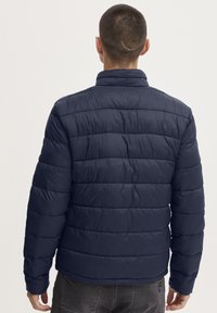 Marinblå pufferjakke med horisontalt quiltede paneler, høy krage og lange ermer. Glatt tekstur, praktisk for kaldt vær.