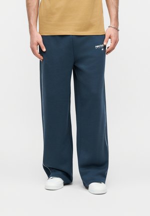 Uomo che indossa pantaloni della tuta New Balance a gamba larga blu navy, sneakers bianche e una camicia color sabbia, in piedi davanti a uno sfondo chiaro e semplice.