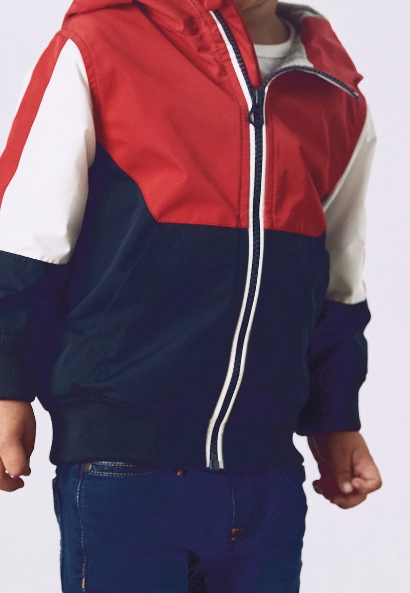 Veste coupe-vent rouge, marine et blanche avec fermeture éclair, poignets élastiques et capuche. Présente un design en blocs de couleur avec une texture lisse.