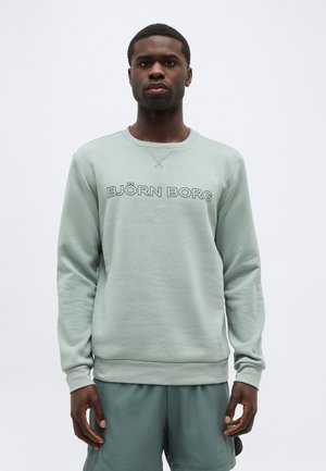 Mand iført en lysegrøn Björn Borg sweatshirt og matchende shorts, stående foran en ensartet lys baggrund.