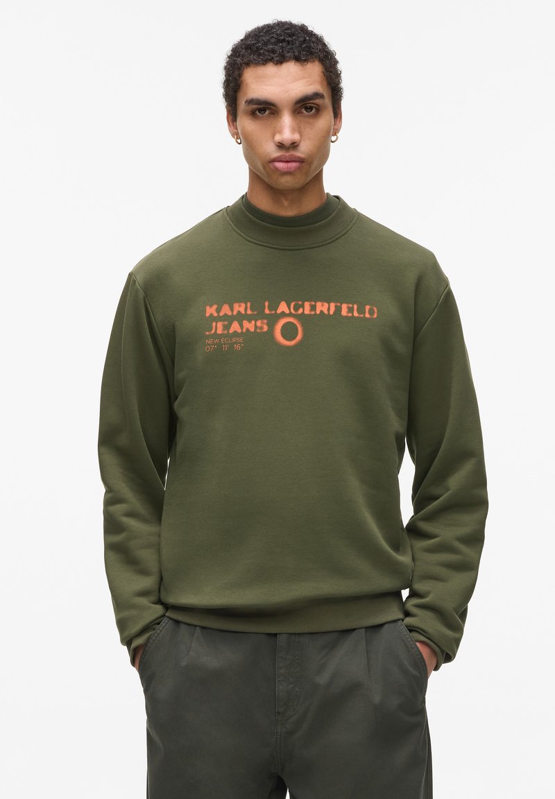 Sudadera verde oliva con cuello alto, con texto impreso en naranja que dice "KARL LAGERFELD JEANS" y un emblema circular. Tejido suave, corte relajado.
