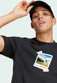 T-shirt en coton noir avec un graphisme rectangulaire imprimé sur la poitrine, représentant un paysage coloré et agrémenté d'accents en ruban.