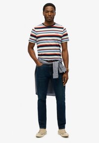 Stribet t-shirt i blå, rød og hvid, parret med mørk denim jeans. En lyseblå skjorte er bundet om livet. Beige sneakers fuldender looket.