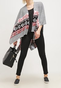 Strikket poncho i sort, hvid og pink med geometriske mønstre, åben front, scalloped kant, parret med en sort top, stramme bukser og en håndtaske.
