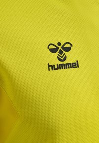 Gult tekstureret stof med en sort stiliseret bi-logo og mærkenavnet "hummel" trykt under logoet.