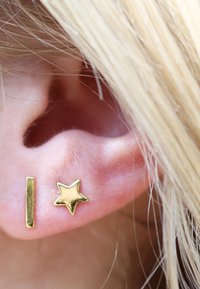 Boucles d'oreilles en or avec une forme rectangulaire verticale et un design en étoile, présentées en gros plan sur une oreille avec des cheveux blonds visibles.