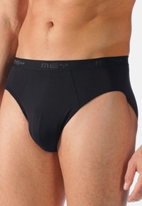Männlicher Oberkörper in schwarzen Slips mit markierter Taille, der Haut von der Taille bis zu den Oberschenkeln vor einem einfarbigen Hintergrund zeigt.