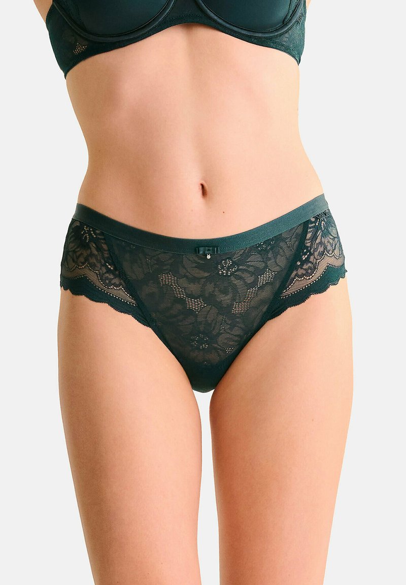 Culotte en dentelle verte avec motif floral, bords festonnés et ceinture lisse. Présente un nœud décoratif au centre frontal.