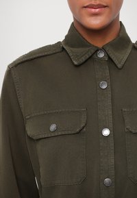 Gros plan d'une personne portant une chemise olive boutonnée avec des poches poitrine à rabat et des boutons-pression argentés.