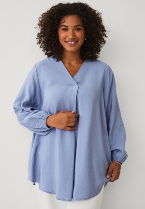 Smiling woman with curly hair wearing a loose, long-sleeve light blue blouse, holding the front near her chest and hem.