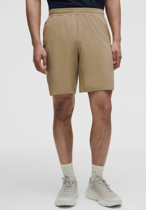 Man met beige short met elastische taille, lichtgekleurde gemêleerde sokken en witte sportschoenen tegen een effen achtergrond.