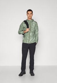 Grøn camo-jakke med lynlås og hætte, parret med sorte bukser. Personen holder en sort rygsæk og har sorte sko på.
