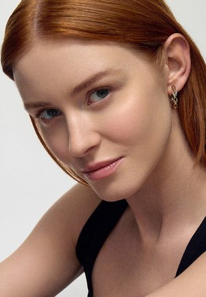 Złote kolczyki w kształcie kółek z połączonymi okrągłymi wzorami są noszone na lewym uchu modelki, prezentując teksturowaną powierzchnię i subtelny blask.