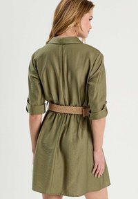 Olijfgroen shirtjurk met korte opgerolde mouwen, cinched taille met een geweven riem en een kraag. Gladde stof met een lichte glans.