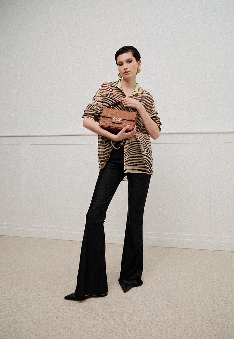 Pantaloni neri a campana e una camicia oversize a motivi marroni e beige, con una borsa marrone in mano. Sfondo neutro e texture del pavimento morbida.