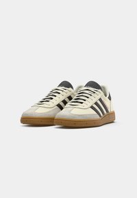 Un par de zapatillas Adidas en tonos crema y gris con rayas negras, suelas marrones y el logo "Spezial" en el lateral, vistas desde la parte frontal izquierda.