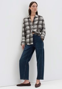 Zwart-wit geruite button-up shirt, semi-gekort, gecombineerd met donkerblauwe Palazzo jeans. Model draagt bruine platte schoenen. Minimalistische achtergrond.