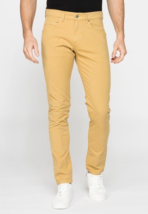 Uomo che indossa pantaloni slim fit giallo senape e sneakers bianche in piedi davanti a uno sfondo chiaro e uniforme.