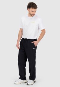 Weißes Baumwoll-T-Shirt mit kleinem gelbem Logo, kombiniert mit schwarzen Hosen mit Kordelzug und weißem Seitenstreifen, getragen mit weißen Sneakers.