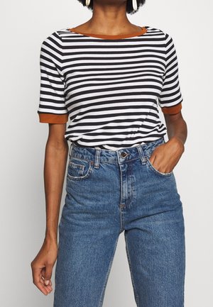 Vrouw die een zwart-wit gestreept T-shirt met korte mouwen en bruine rand draagt, gecombineerd met een blauwe jeans met hoge taille, met één hand in de zak.