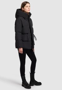 Zwarte pufferjack met een hoge kraag en een trekkoord in de taille, met een gladde, matte textuur en een rits aan de voorkant, gedragen met zwarte leggings en laarzen.