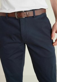 Homme portant un pantalon bleu foncé avec une ceinture en cuir marron et une chemise blanche, la main reposant près de la poche.