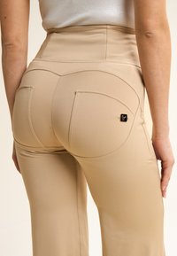 Pantaloni beige a vita alta con una silhouette attillata, dotati di tasche posteriori e un piccolo logo decorativo. Tessuto liscio e design sartoriale.