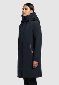 Mørkeblå lang frakke med høj krave og hætte, der har sidedel med lynlås og glat, tekstureret stof.