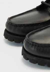 Chaussures en cuir noir avec un bout arrondi, couture visible et une semelle en caoutchouc texturée présentant un motif robuste.